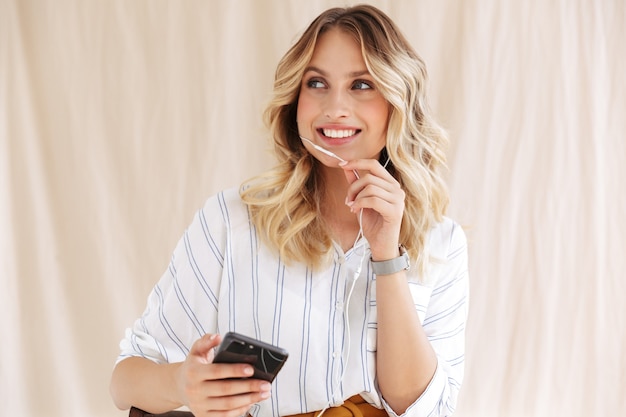 uwodzicielska elegancka blondynka w słuchawkach uśmiechająca się i trzymająca smartfona