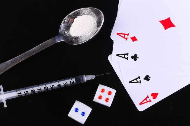 Zdjęcie uzależnienie od narkotyków i hazardu. łyżka z proszkiem narkotykowym, strzykawką, kostką do gry i czterema asami na białym tle na czarnym tle.