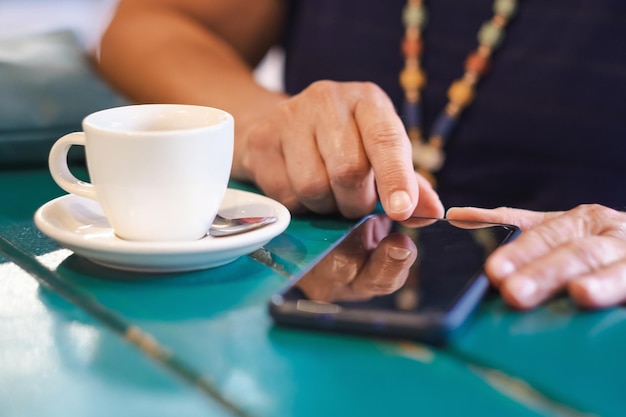 Uzależniona dorosła dojrzała kaukaska kobieta siedzi przy stole z białą filiżanką kawy za pomocą telefonu komórkowego