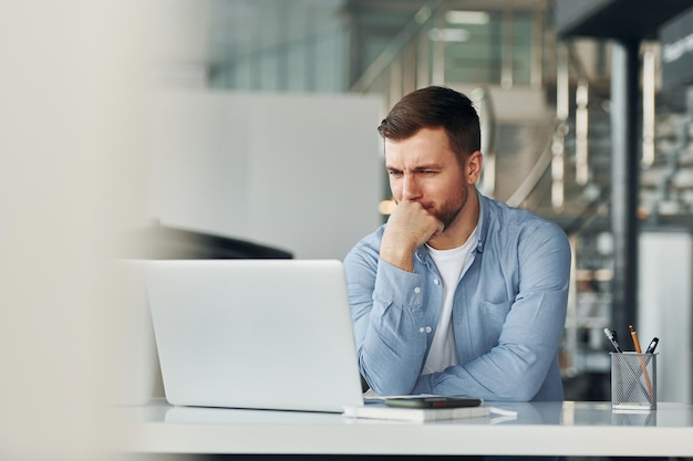 Używa laptopa Młody mężczyzna w białej koszuli i niebieskiej kurtce pracuje w pomieszczeniu