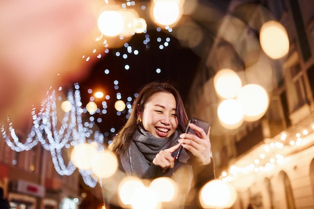 Używa telefonu Śliczna i szczęśliwa młoda azjatycka kobieta na zewnątrz w mieście świętuje Nowy Rok