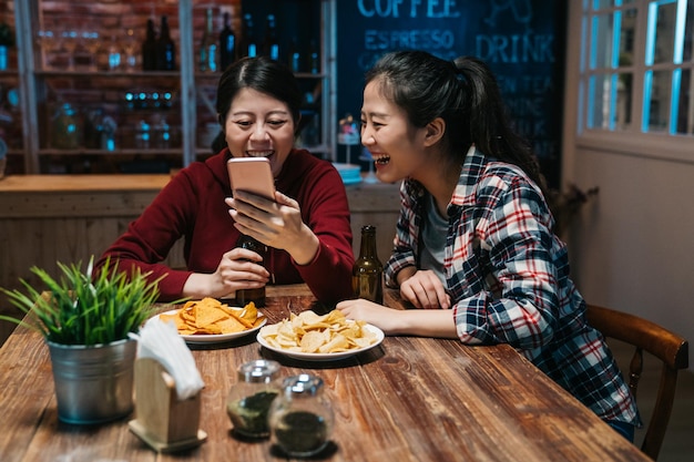 w barze jednonocne świętowanie w toku piękni młodzi ludzie pokazują ekran telefonu komórkowego, mając świetną zabawę. dwie najlepsze przyjaciółki Azjatki, śmiejące się, przeglądające media społecznościowe, piją piwny pub.