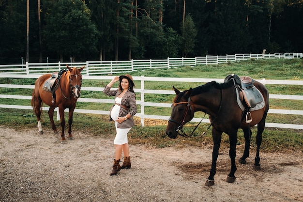 W ciąży dziewczyna z dużym brzuchem w kapeluszu obok koni w pobliżu padoku w przyrodzie.