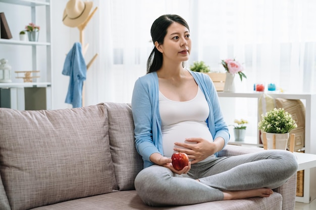 W ciąży piękna dziewczyna azjatyckich trzymać czerwone jabłko. Atrakcyjna mama czuje się zrelaksowana i szczęśliwa. urocza kobieta ma dobre zdrowie dba o siebie jedząc zdrowe jedzenie siedząc na kanapie w domu