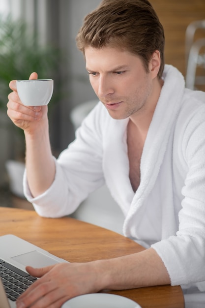 W Internecie. Przystojny mężczyzna w białym szlafroku siedzi przy laptopie i spędza czas w sieci