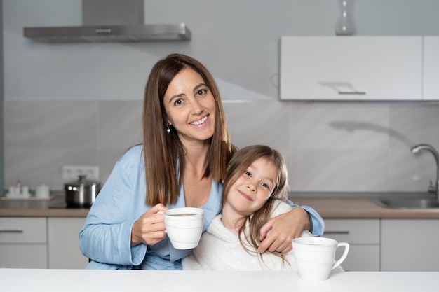 W kadrze wesoła mama i córka siedzą razem przy kuchennym stole pijąc rano gorącą herbatę, prowadząc ze sobą miłą rozmowę