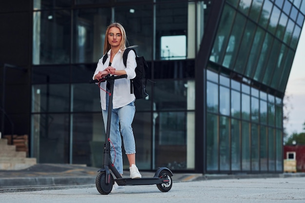 W pobliżu budynku biznesowego Piękna blondynka w zwykłych ubraniach jeździ na skuterze elektrycznym na zewnątrz w słoneczny dzień