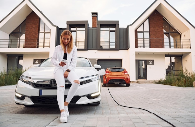 W pobliżu domu Młoda kobieta w białym ubraniu jest ze swoim samochodem elektrycznym w ciągu dnia