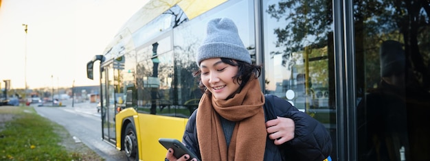 W pobliżu znajduje się zdjęcie studentki oczekującej na rozkład jazdy transportu publicznego w aplikacji na smartfona