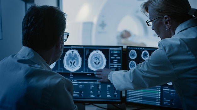 W pokoju kontrolnym lekarz i radiolog omawiają diagnozę podczas obserwacji zabiegu, a monitory pokazują wyniki skanowania mózgu w tle Pacjent przechodzi zabieg MRI lub CT