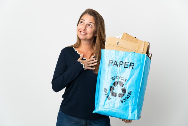W średnim wieku Brazylijska kobieta trzyma worek recyklingu pełnego papieru do recyklingu na izolowanej ścianie patrząc w górę uśmiechając się