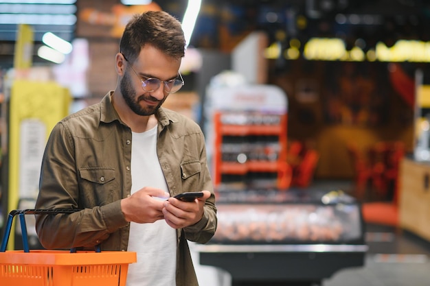 W supermarkecie przystojniak używa smartfona i patrzy na wartość odżywczą konserwów Stoi z koszykiem na zakupy w sekcji konserw