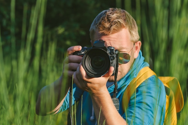 W zbliżeniu fotograf trzyma w rękach profesjonalną kamerę foto-wideo. Na tle zieleni pięknej przyrody.