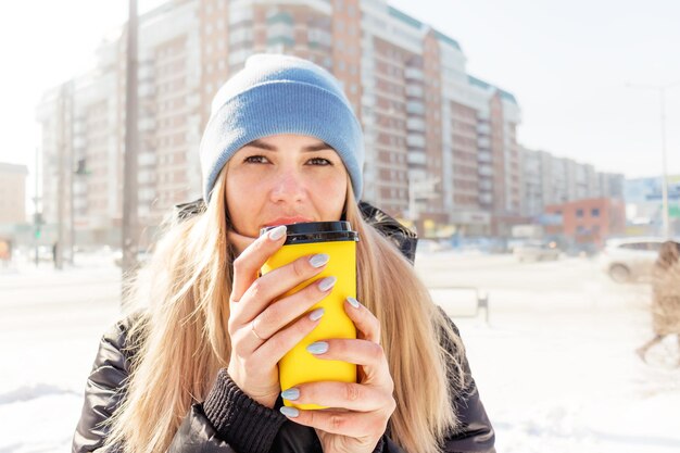 Zdjęcie w zimowy poranek młoda kobieta trzyma żółty papierowy kubek z kawą i rozgrzewa ręce kawą.