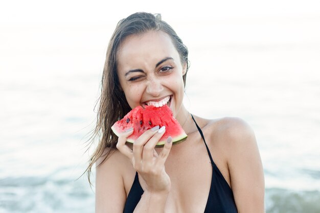 Zdjęcie wakacje, resort, koncepcja turystyki - letnie wakacje - młoda dziewczyna jedzenie świeżego arbuza na piaszczystej plaży. młoda piękna kobieta zjada arbuza na plaży w upalny letni dzień.