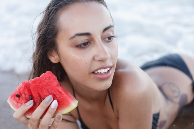 Wakacje, resort, koncepcja turystyki - letnie wakacje - młoda dziewczyna jedzenie świeżego arbuza na piaszczystej plaży. młoda piękna kobieta zjada arbuza na plaży w upalny letni dzień.