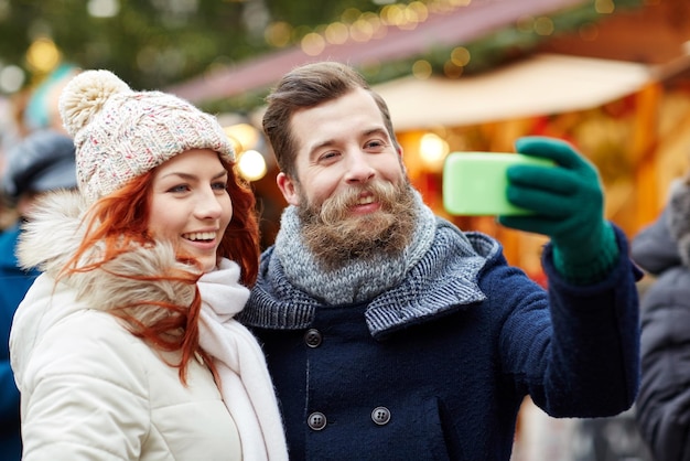 wakacje, zima, święta, technologia i koncepcja ludzi - szczęśliwa para turystów w ciepłych ubraniach robi selfie ze smartfonem na starym mieście