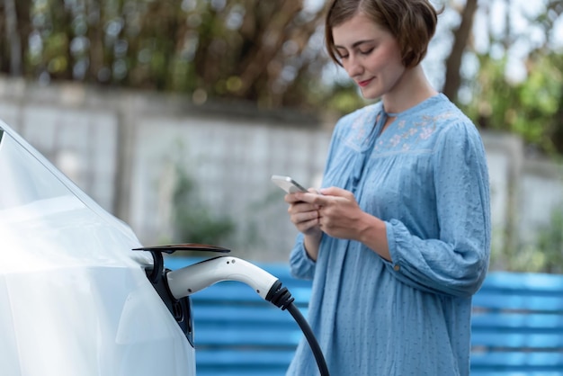 Wakacyjna wycieczka samochodowa wśród zielonej wiejskiej przyrody z piękną młodą kobietą sprawdzającą stan baterii na smartfonie podczas ładowania pojazdu elektrycznego Ekologiczne podróżowanie samochodem elektrycznymPerpetual