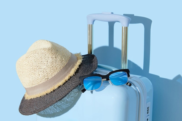 Walizka z okularami przeciwsłonecznymi i kapeluszem na pastelowym błękitnym tle Koncepcja podróży Minimalistyczny styl