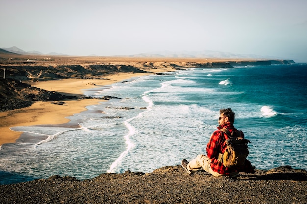 Wanderlust and travel people concept z hipsterskim mężczyzną siedzącym na skałach na klifie ze wspaniałym pięknym widokiem na naturalną nieodkrytą plażę