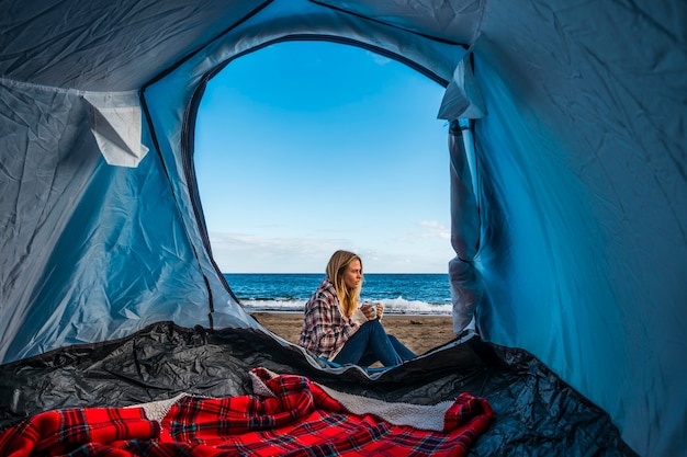 Wanderlust i alternatywny wakacyjny styl życia dla blond ładnej dziewczyny siedzącej na zewnątrz namiotu biwakującego na piasku na plaży przed falami oceanu