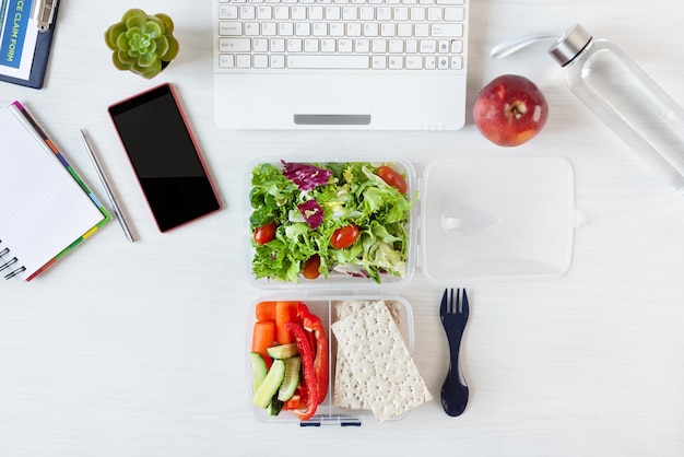 Warzywa i świeża zielona sałatka w pudełku na lunch na biurku z laptopem i innymi artykułami biurowymi