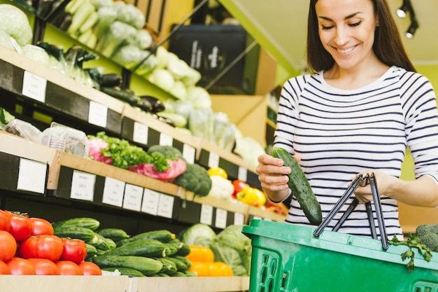 Warzywa na letnią sałatkę Młoda kobieta w luźnych ciuchach wybiera ogórka z półki w supermarkecie w dziale owoców i warzyw