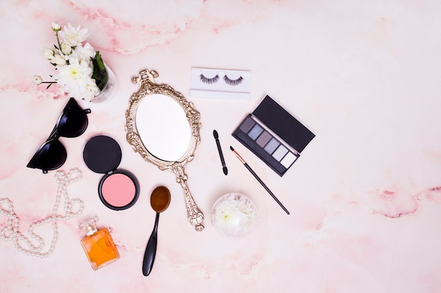 Wazon; okulary słoneczne; naszyjnik; ręczne lusterko; kompaktowy puder do twarzy; pędzel do makijażu; rzęsy i paleta cieni do powiek na różowym tle