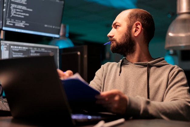 Web developer analizujący kod źródłowy wraz z dokumentami i komputerem, robiący notatki i pracujący nad tworzeniem stron internetowych. Męski programista porównujący pliki z danymi serwera online po godzinach.