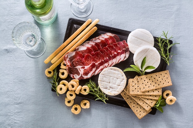 Wędliny I Sery Podawane Są Na Tacy Na Stole Z Białym Winem, Krakersami, Grissini I Taralli Z Aromatycznymi Ziołami Na Niebieskim Obrusie Lnianym.