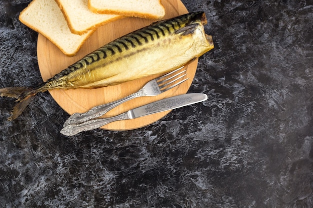 Wędzona makrela bez głowy z widelcem nożem deska do krojenia chleb na czarnym tle copyspace widok z góry