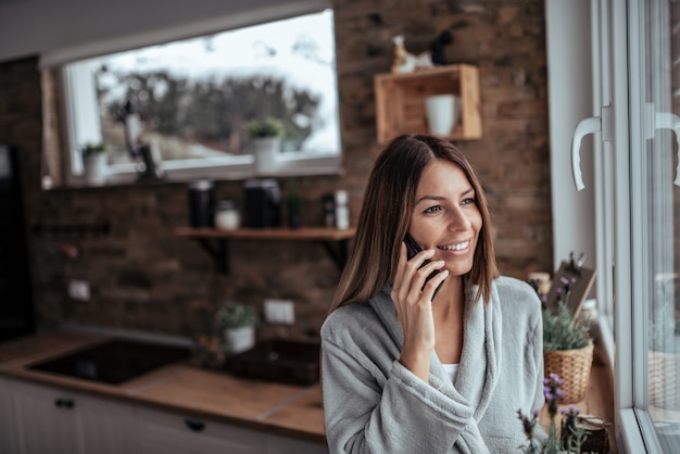 Weekendowe poranki. Piękna brunetka patrzeje przez okno i opowiada na mądrze telefonie na zima dniu w cosy kuchni.