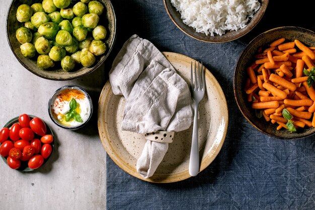 Wegański stół obiadowy z ryżem i warzywami pieczonymi brukselką, marchewką baby, pomidorkami koktajlowymi w ceramicznych miskach i pustym talerzem gości na niebieskim lnianym obrusie na szarym tle. Płaski układanie.