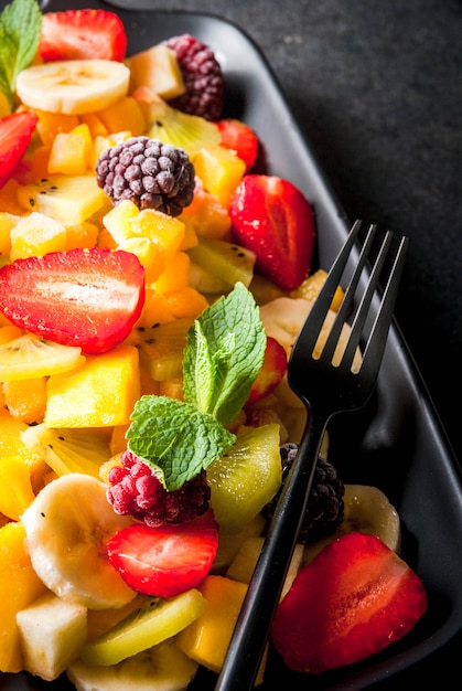 Wegańskie potrawy dietetyczne. Witaminy Deser. Lato. Sałatka ze świeżych owoców mango, brzoskwini, jabłka, banana, kiwi, truskawki, jeżyn. Na czarnej płycie ceramicznej miejsce na czarny kamienny stół