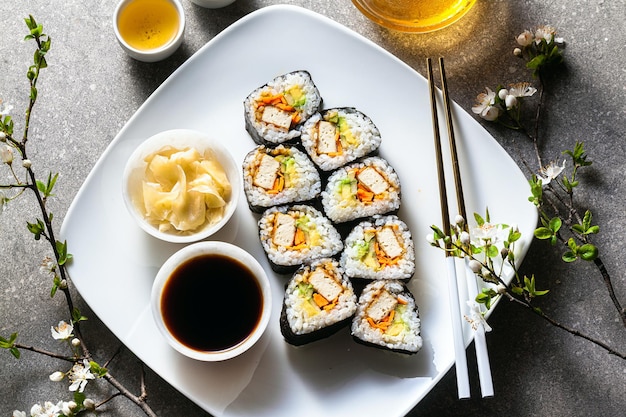 Wegańskie sushi z tofu w sosie General Tso awokado i świeżymi warzywami na stole z gałęziami kwitnącego drzewa i zieloną herbatą w szklanym czajniczku