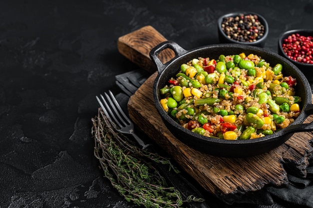 Wegetariańska smażona sałatka z komosy ryżowej z edamame, kukurydzą, marchewką, pomidorem, papryką, cebulą na patelni. Czarne tło. Widok z góry. Skopiuj miejsce.