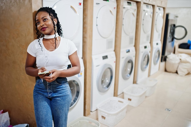 Wesoła afroamerykanka w pobliżu pralki słuchająca muzyki przez słuchawki z telefonu komórkowego w samoobsługowej pralni