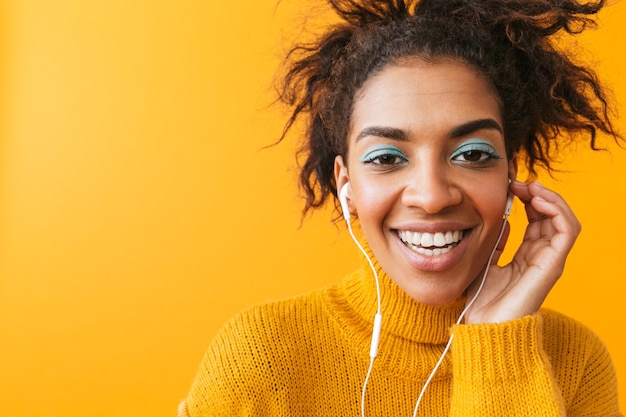 Wesoła afrykańska kobieta ubrana w sweter stojący na białym tle, słuchanie muzyki przez słuchawki