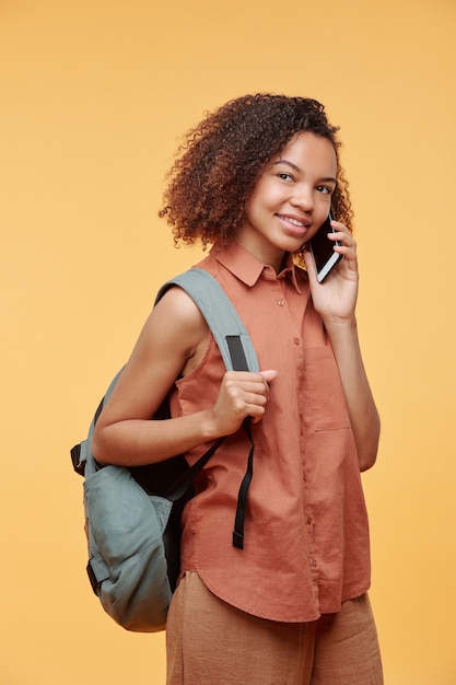 Wesoła atrakcyjna dziewczyna afroamerykańskiego studenta w casual shirt trzymając tornister i dzwoniąc do szkolnego przyjaciela przez telefon na żółtym tle