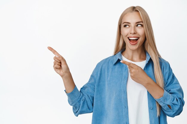 Wesoła, atrakcyjna kobieta wskazująca palcami w lewo, patrząca na reklamę, pokazująca ogłoszenie, stojąca na białym