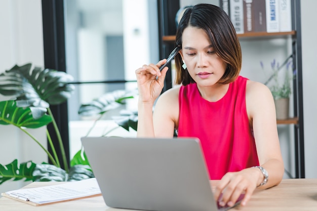 Wesoła Azjatycka bizneswoman w średnim wieku w swobodnej sukience dorywczo pracująca w domu, sprawdzająca pocztę e-mail na komputerze przenośnym, pisząca na papierze z dokumentami księgowymi. Biznesowe zdjęcie stockowe