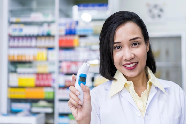 Wesoła azjatycka farmaceuta chemik kobieta z cyfrowym termometrem stojąca w aptece apteka