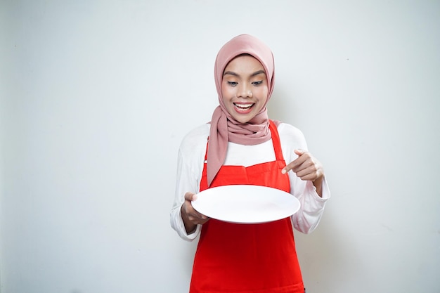 Wesoła azjatycka muzułmańska kobieta w czerwonym fartuchu wskazująca na pusty talerz Reklama żywności Koncepcja gotowania