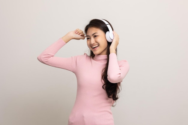 Wesoła azjatycka nastolatka słucha muzyki z białymi słuchawkami na białym tle Piękna młoda kobieta w różowej koszuli dłoni dotyka bezprzewodowych słuchawek bawiąc się muzyką w pokoju