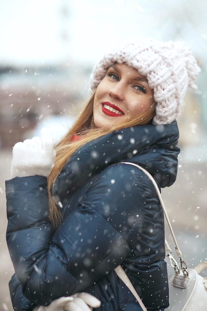 Wesoła blond kobieta nosi czapkę z dzianiny spaceru po mieście podczas opadów śniegu. Miejsce na tekst