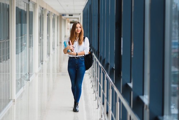 Wesoła brunetka dziewczyna studentka z czarnym plecakiem trzyma książki w nowoczesnym budynku