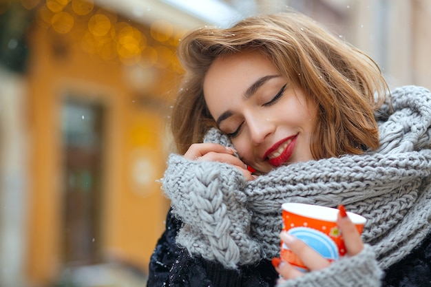 Wesoła brunetka kobieta nosi szalik z dzianiny i cieszy się śnieżną pogodą i pije kawę. Pusta przestrzeń