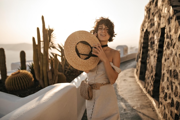Wesoła brunetka w beżowej sukience ze słomkowym kapeluszem i małą torebką porusza się po ścieżce z kaktusami i pięknym widokiem na morze