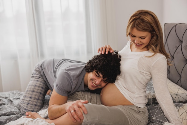 Zdjęcie wesoła ciężarna para bawiąca się podczas relaksu w sypialni. szczęśliwy ojciec słuchający bicia serca dziecka.