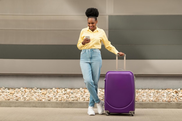 Wesoła czarna kobieta z walizką podróżną za pomocą SMS-ów na zewnątrz